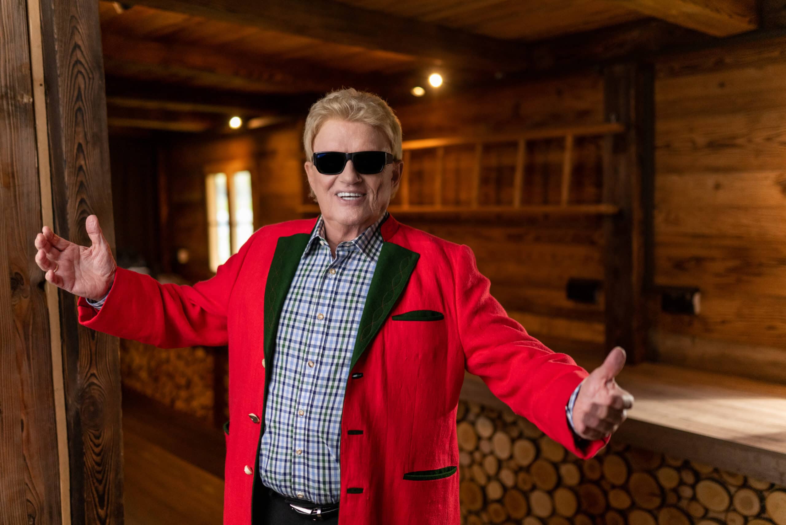 Im roten Trachtenjanker und mit dunkler Sonnenbrille präsentiert sich ein Heino in rustikaler Umgebung – Foto: Helmut Werner Management.